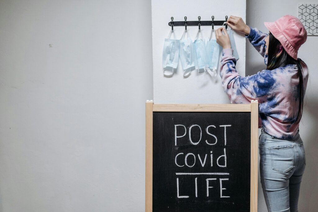 A person hangs masks near a chalkboard with 'Post Covid Life' written, symbolizing the transition to normalcy.